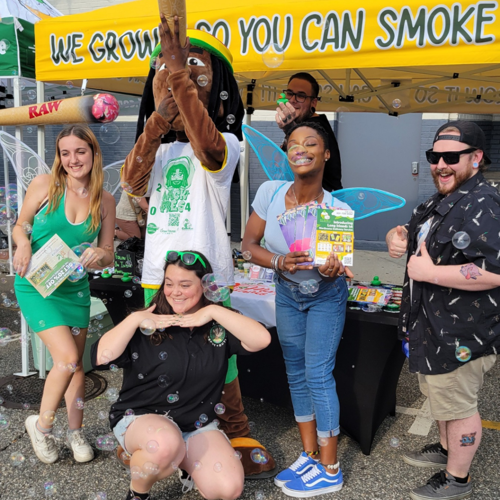Group of people celebrating at an EZ Bud event, featuring a mascot holding a large joint, colorful promotional materials, and bubbles, with a vibrant tent backdrop displaying "WE GROW SO YOU CAN SMOKE."